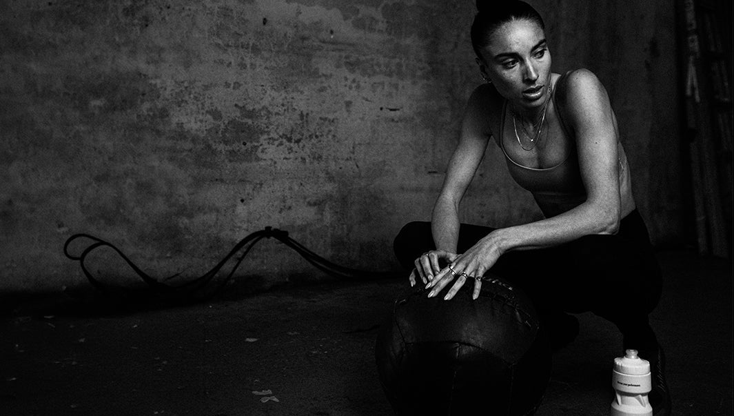 A black and white photograph of a woman crouching next to a medicine ball and a water bottle in a dark, gritty room with a textured concrete wall.