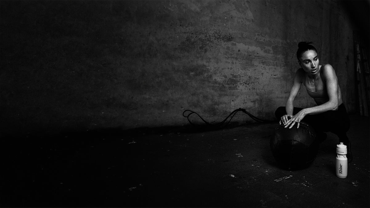 A black and white photograph of a woman crouching next to a medicine ball and a water bottle in a dark, gritty room with a textured concrete wall.